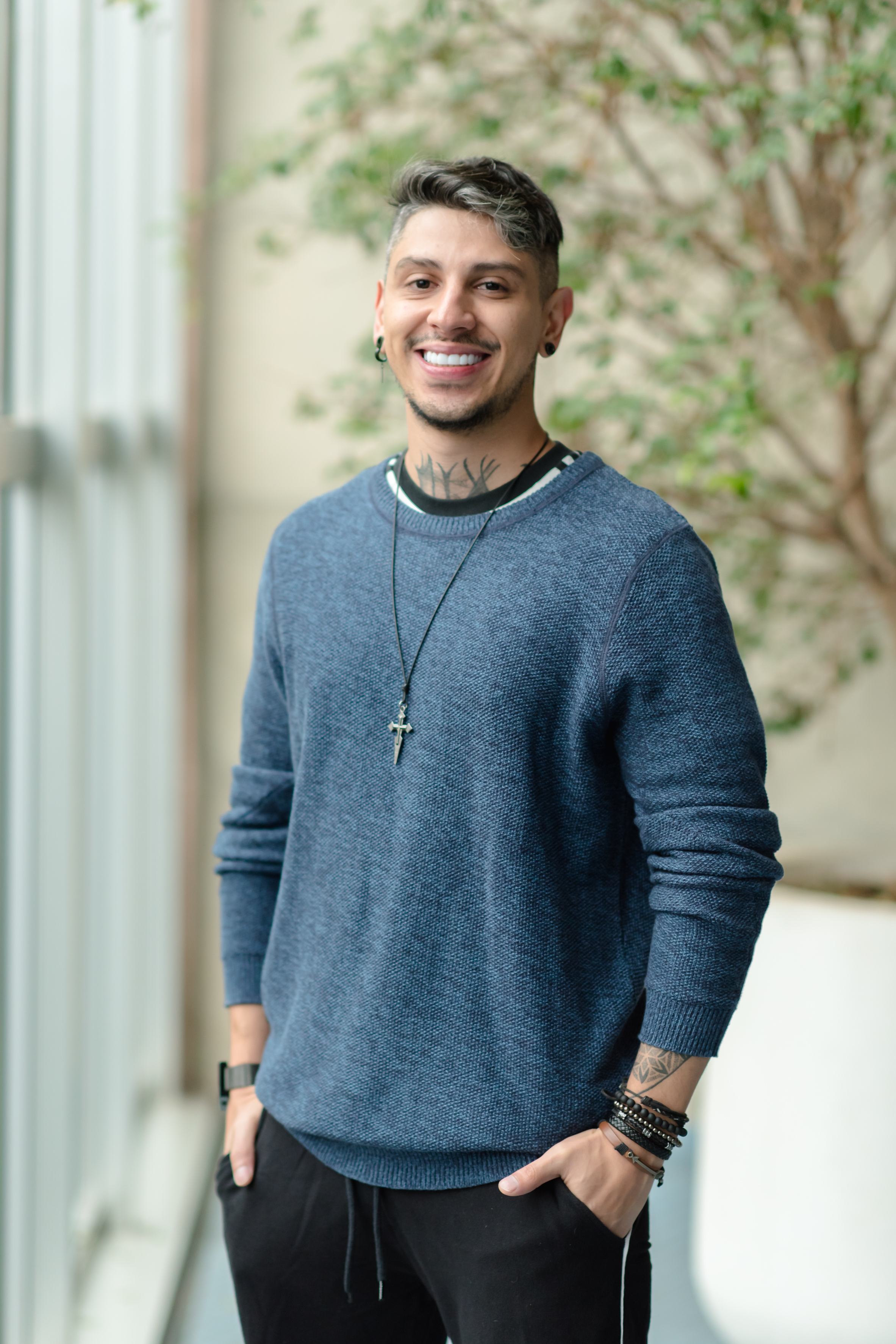 A imagem mostra um homem sorridente em pé em um ambiente interno com vegetação ao fundo. Ele tem cabelo curto e grisalho, usa um brinco na orelha esquerda e tem tatuagens visíveis no pescoço e no braço. Está vestido com uma camiseta de mangas compridas em tom azul e uma calça preta. Usa um colar com um pingente de cruz e várias pulseiras pretas no pulso. Ele parece estar feliz e relaxado, com uma expressão amigável e acolhedora.